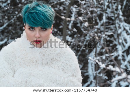 A nice girl with blue hair wrapped herself in a white fur coat in a snow-covered park. Rubbing against the background of a bush