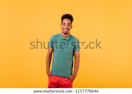 Indoor portrait of smiling black man standing with hands in pockets. Studio shot of blithesome african guy isolated on yellow background.