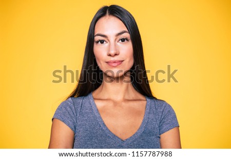 Young attractive cute brunette woman in gray T-shirt is posing and looking on camera isolated over yellow wall