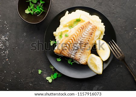 Fried cod white fish with mashed potatoes and lemon slices on a black plate and a dark background. Top View