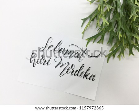 Selamat hari merdeka (happy independence day) hand lettered on a piece of white paper with black ink against a white background with green plant on the top 
