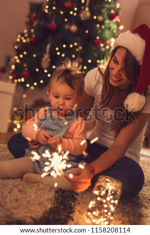 Beautiful young mother and daughter sitting on the floor next to a Christmas tree, playing with sparklers and christmas lights