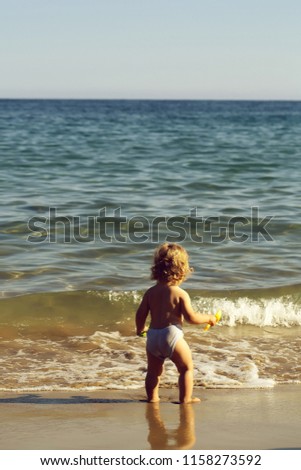 Small curious blonde child boy standing on sea coast beach with wavy water sunny twilight outdoor playing on natural background, vertical picture