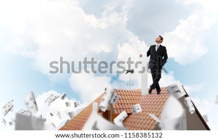 Conceptual image of young businessman in suit looking upside and holding big white arrow in hand while standing on brick roof among flying papers and cloudy skyscape view on background.