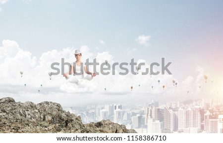 Young woman in white clothing keeping eyes closed and looking concentrated while meditating on cloud in the air with city view on background.