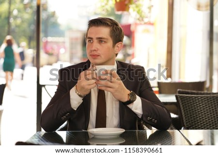 a young man in a black business suit sits in a street cafe at the table and enjoys a cappuccino with foam
