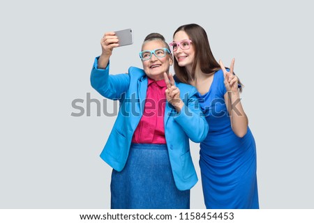 happy grandmother with granddaughter in blue dress or suit toothy smiling, standing, posing and making selfie and peace sign. Relations in the family. indoor, studio shot, isolated on gray background