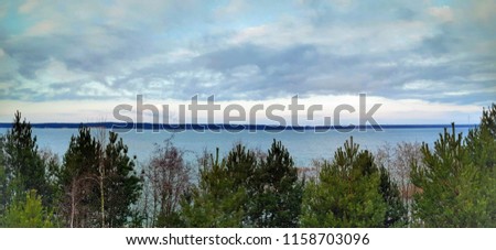 Light blue lake with green and brown forest trees and buskes in front of it, lake is flooded sand quarry in Germany, cloudy sky and skyline in the background