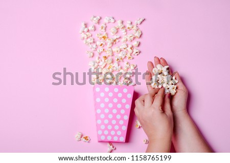 Paper box with popcorn on pink background with female hands. Top view, copy space