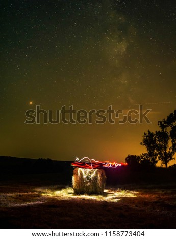 The Milky Way shines bright over a straw bales. Long exposure with light painting. Red and white long exposure colors around the straw bales and the milk way in background.