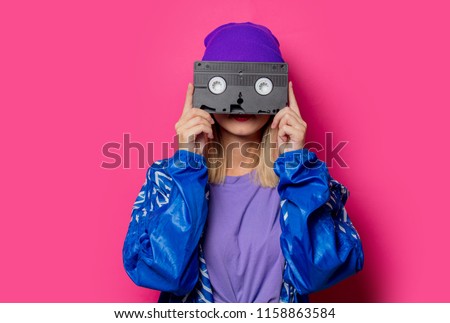 Young blonde girl in 90s sports jacket and VHS cassette on pink background. Royalty-Free Stock Photo #1158863584