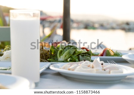Restaurant at sea shore against blue water and Yacht Port of Aegean coast on Cesme. Luxury table with plate of fish appetizers, cheese, meze, salad, octopus, raki and ouzo. Copy space for text area.