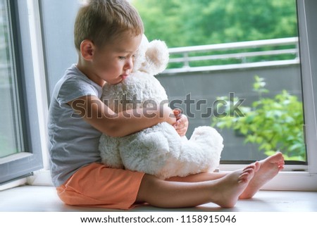 boy of two years sitting by the window and hugs a toy Bunny. rainy weather, waiting for dad to come home from work