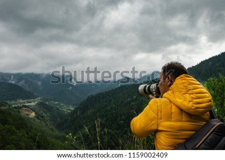 A man in a vibrant yellow coat and with a backpack is taking picture of the misty mountains and valley on a rainy cloudy day