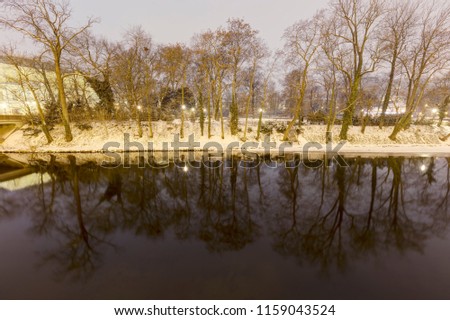 HDR winter photo of trees on the bank of a river with snow covering the ground and trees reflecting in still water.