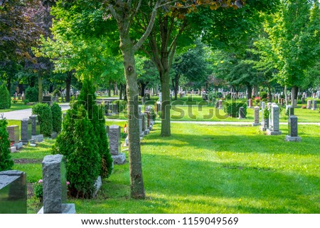 A beautiful graveyard marked with rows of headstones and lined with trees, flowers and walking paths, providing a peaceful setting for visitors to visit departed beloved friends and family members. Royalty-Free Stock Photo #1159049569