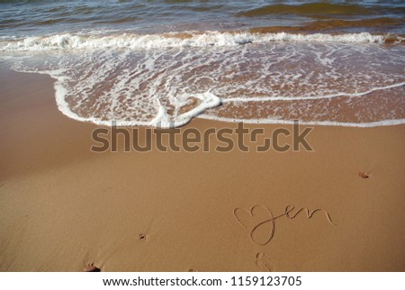 A  message "Zen" beautifully handwritten in a cursive style on the red sand, on a beach with the blue ocean and a wave in the upper part, Prince Edward Island, Canada