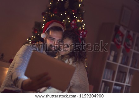 Couple in love sitting next to a Christmas tree, wearing Santa's hats and having fun looking at old photos on a tablet computer.