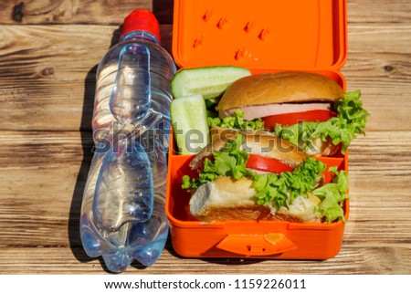Bottle of water and school lunch box with two homemade burgers and fresh cucumbers on wooden table