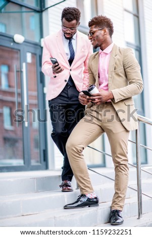 stylish guy in white suit showing his photos on the smart phone to his colleague while they standing on the stairs