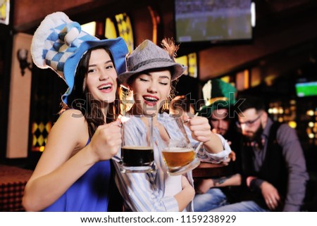 friends - boys and girls in Bavarian hats drinking beer on the background of the bar during the celebration of Oktoberfest or St. Patrick's Day