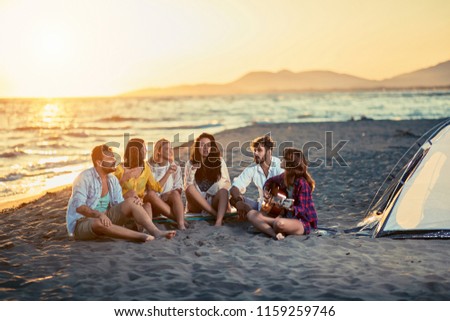 Group of happy friends with guitar at beach. friends relaxing on sand at beach with guitar and singing on summer sunset
