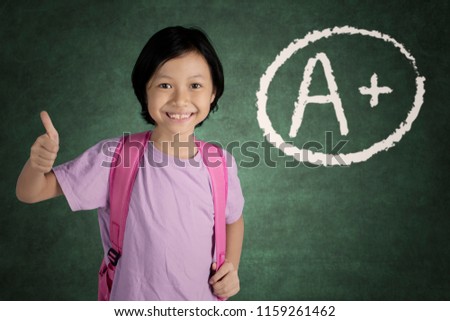 Picture of cute schoolgirl looks happy while getting grade A plus while showing her thumb in the classroom