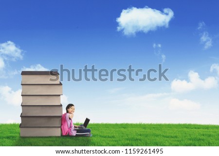 Image of happy schoolgirl showing thumb up while using a laptop and sitting near a pile of books in the meadow