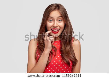 Horizontal shot of attractive young European female wears red lipstick, has joyful expression, smiles broadly, dressed in summer red polka dot dress, being in good mood as has date with boyfriend