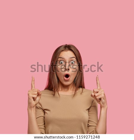 Stunned Caucasian female has shocked expression, points upwards with both index fingers, cant believe in something, shows blank space above head, has appealing appearance. People and reaction