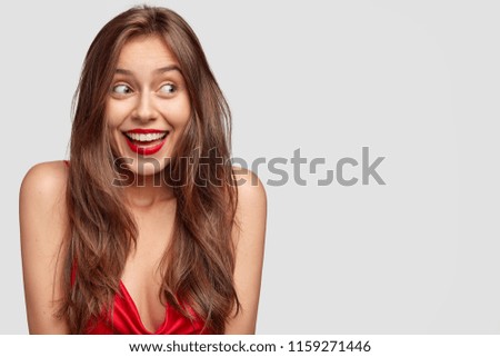 Attractive young female with long dark straight hair, has happy expression, red lips, dressed casually, stands against white background with copy space for your advertisement or promotional text