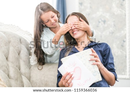 Charming little girl is giving her beautiful young mom a present, woman is smiling while sitting on couch at home. daughter closes her eyes to mom