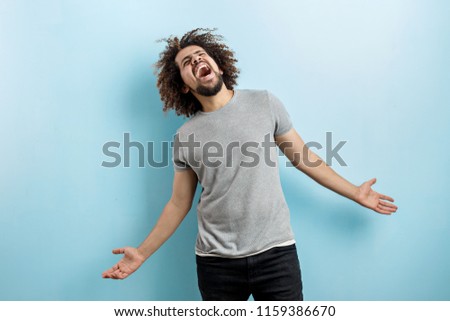 A curly-headed handsome man wearing a gray T-shirt is standing and laughing hard with his hands widly spread, looking upwards over the blue background. Happiness and joy.