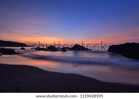 The curved waves in front of the beach rock reflect the sunrise, sunset, blue sky and orange clouds.