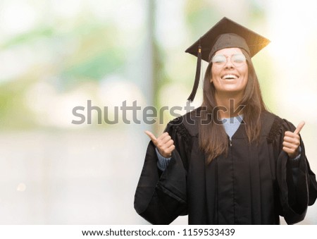 Young hispanic woman wearing graduated cap and uniform success sign doing positive gesture with hand, thumbs up smiling and happy. Looking at the camera with cheerful expression, winner gesture.