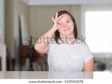Down syndrome woman at home with happy face smiling doing ok sign with hand on eye looking through fingers