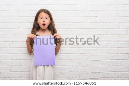 Young hispanic kid over white brick wall holding pink paper sheet scared in shock with a surprise face, afraid and excited with fear expression