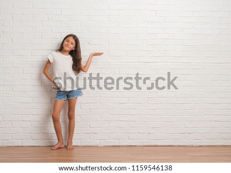 Young hispanic kid stading over white brick wall smiling cheerful presenting and pointing with palm of hand looking at the camera.
