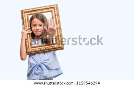 Brunette hispanic girl holding vintage art frame with open hand doing stop sign with serious and confident expression, defense gesture