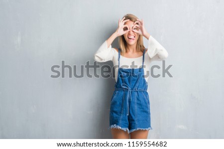 Beautiful young woman standing over grunge grey wall doing ok gesture like binoculars sticking tongue out, eyes looking through fingers. Crazy expression.
