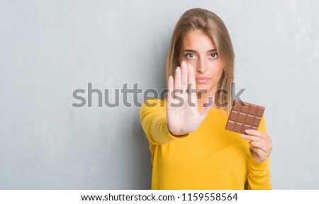 Beautiful young woman over grunge grey wall eating chocolate bar with open hand doing stop sign with serious and confident expression, defense gesture