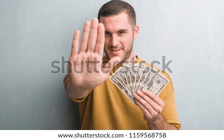 Young caucasian man over grey grunge wall holding dollars with open hand doing stop sign with serious and confident expression, defense gesture