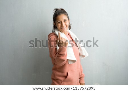 Young sporty indian woman against a wall inviting to come, confident and smiling making a gesture with hand, being positive and friendly. Holding a towel after excercising.
