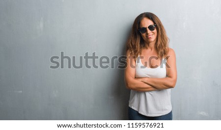Middle age hispanic woman over grey wall wearing sunglasses happy face smiling with crossed arms looking at the camera. Positive person.