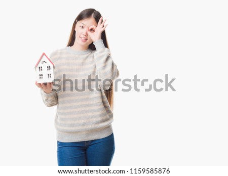Young Chinese real state agent woman over isolated background holding house with happy face smiling doing ok sign with hand on eye looking through fingers