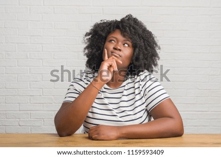Young african american woman sitting on the table at home with hand on chin thinking about question, pensive expression. Smiling with thoughtful face. Doubt concept.