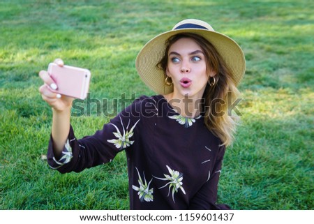 Young beautiful woman in trendy hat making funny faces on her phone on a green grass in a park