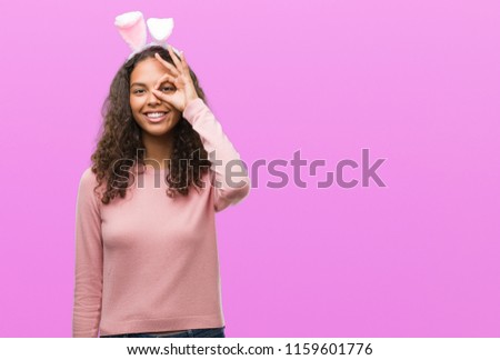 Young hispanic woman wearing easter bunny ears with happy face smiling doing ok sign with hand on eye looking through fingers