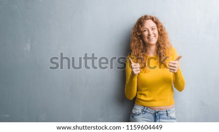 Young redhead woman over grey grunge wall success sign doing positive gesture with hand, thumbs up smiling and happy. Looking at the camera with cheerful expression, winner gesture.