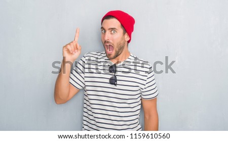 Handsome young hipster man over grey grunge wall wearing navy t-shirt and wool cap pointing finger up with successful idea. Exited and happy. Number one.